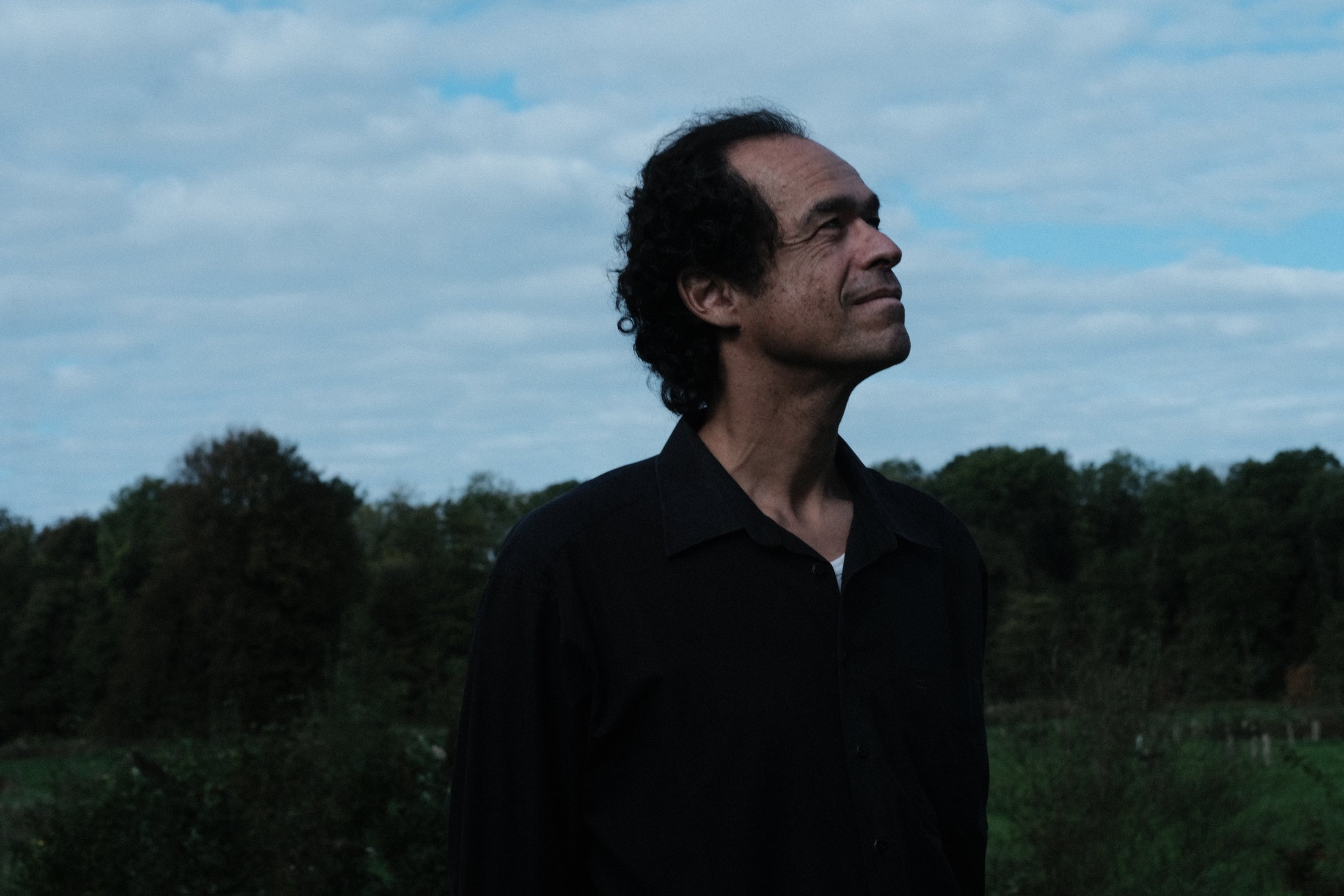 Jérôme Inen in a black shirt, looking up towards a cloudy sky beside a line of trees
